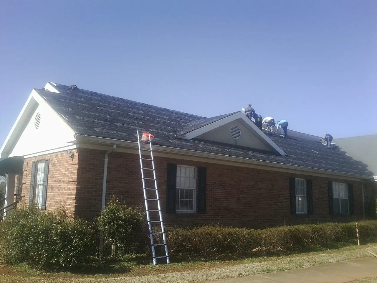 Crew tearing shingles off a brick ranch roof with a ladder set — Emergency Roof Repair work by our crew in Lena