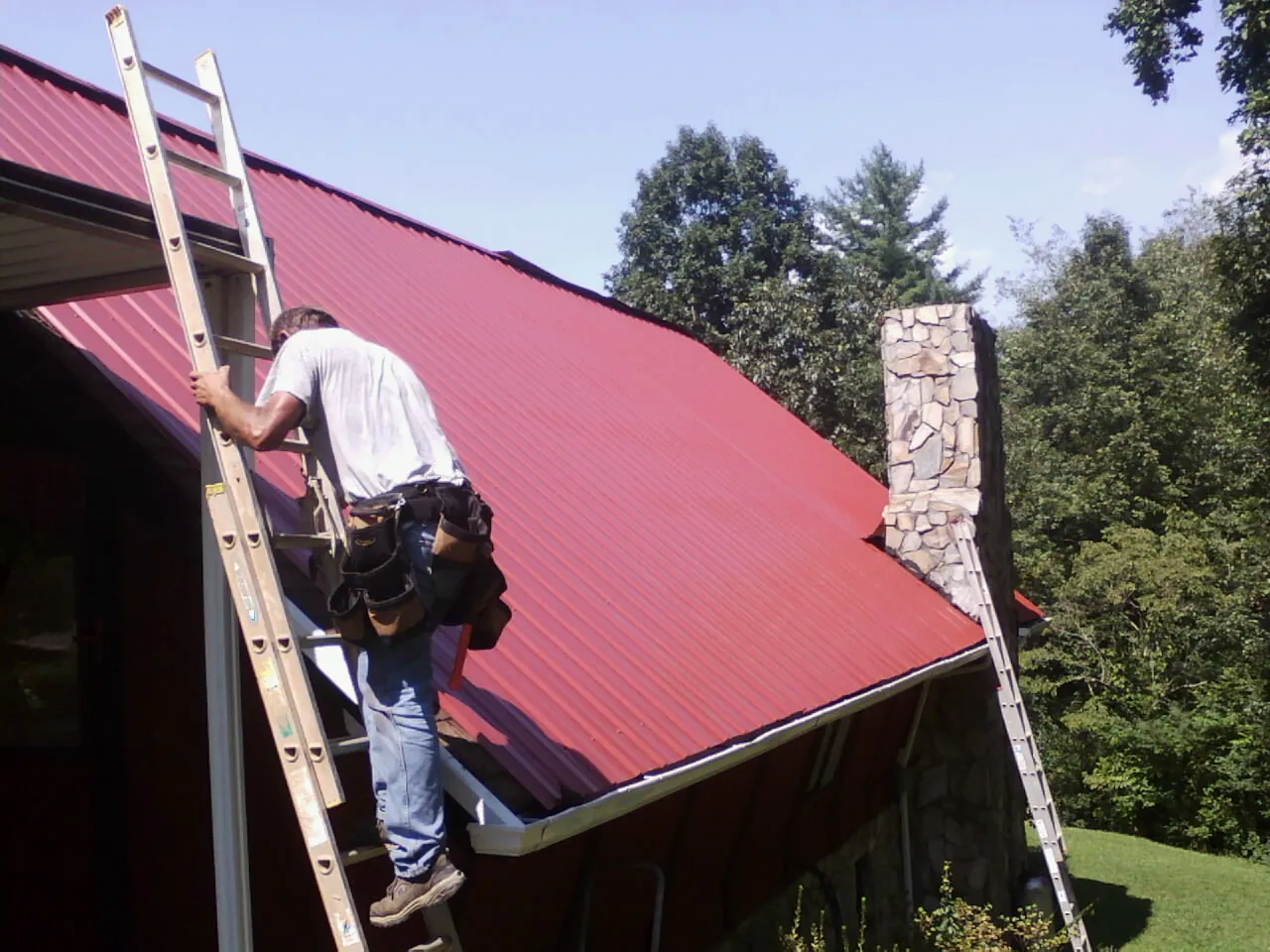 Roofer on a ladder beside a red metal roof and stone chimney — Soffit & Fascia Repair by Roofing Brothers in Lena